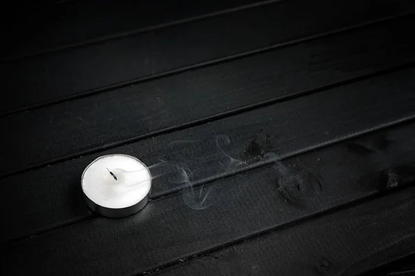 A small white candle on a dark wooden table. Smoke from a blown-out candle. Black tabletop boards. decorative candle.