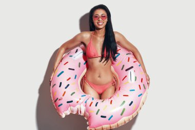 Bikinili çekici genç bir kadın gri arka planda dururken kameraya bakıyor ve gülümsüyor.