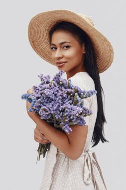 Çekici, şapkalı, gülümseyen ve gri arka planda kameraya bakan genç bir kadın.