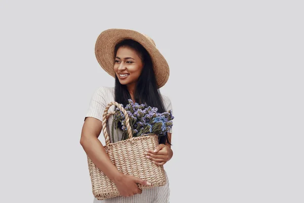 Atractiva joven con sombrero sonriendo y llevando canasta con flores mientras está de pie sobre fondo gris —  Fotos de Stock