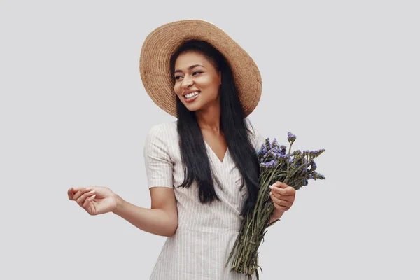 Atractiva joven con sombrero sonriendo y llevando ramo mientras está de pie sobre fondo gris —  Fotos de Stock