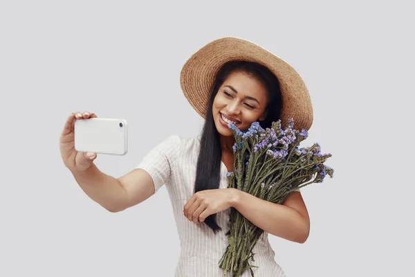 Attractive young woman in hat smiling and taking selfie while standing against grey background