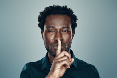 Yakışıklı Afrikalı genç adam kameraya bakıyor ve parmağını dudaklarının üzerinde tutuyor gri arka planda duruyor.