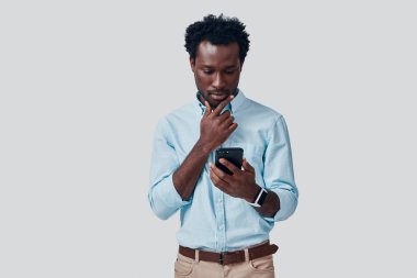Yakışıklı genç Afrikalı adam gri arka planda dururken akıllı telefon kullanıyor.