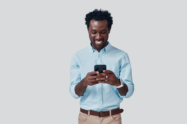 Beau jeune homme africain utilisant un téléphone intelligent et souriant tout en se tenant debout sur fond gris — Photo