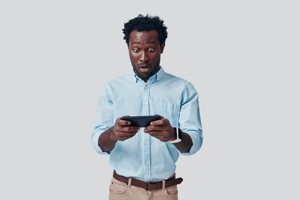 Όμορφος νεαρός Αφρικανός παίζει κινητό παιχνίδι, ενώ στέκεται πάνω από το γκρι φόντο — Φωτογραφία Αρχείου