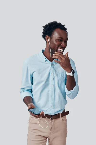 Όμορφος νεαρός Αφρικανός που ακούει μουσική και χαμογελάει ενώ στέκεται στο γκρίζο φόντο — Φωτογραφία Αρχείου