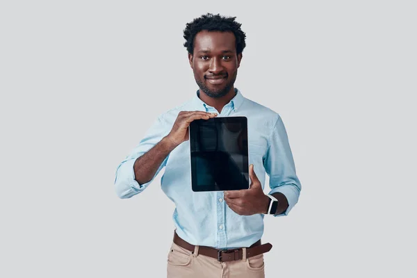 Όμορφος νεαρός Αφρικανός που δείχνει το χώρο των αντιγράφων στο ψηφιακό tablet και χαμογελά ενώ στέκεται πάνω σε γκρι φόντο — Φωτογραφία Αρχείου