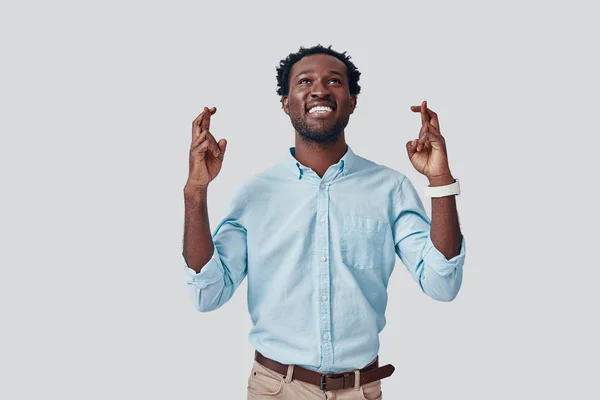 Όμορφος νεαρός Αφρικανός σταυρώνει τα δάχτυλα και χαμογελά ενώ στέκεται πάνω σε γκρι φόντο — Φωτογραφία Αρχείου