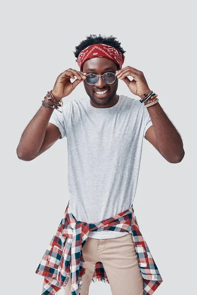 Όμορφος νεαρός Αφρικανός προσαρμόζει τα γυαλιά του και κοιτάζει την κάμερα ενώ στέκεται στο γκρίζο φόντο — Φωτογραφία Αρχείου