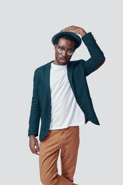 Όμορφος νεαρός Αφρικανός προσαρμόζει το καπέλο και κοιτάζει την κάμερα ενώ στέκεται στο γκρίζο φόντο — Φωτογραφία Αρχείου