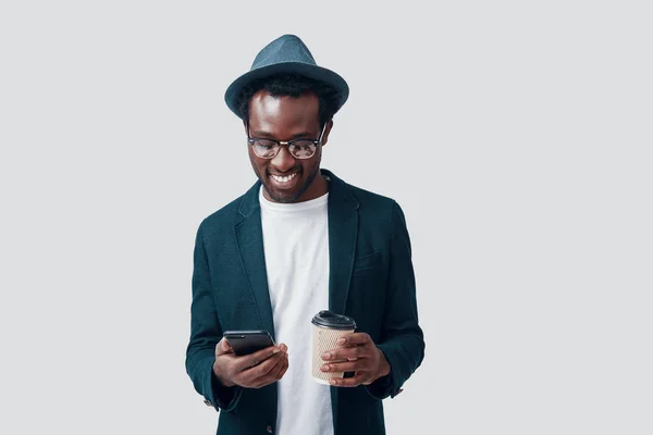 Beau jeune homme africain utilisant un téléphone intelligent et tenant une tasse jetable tout en se tenant debout sur fond gris — Photo