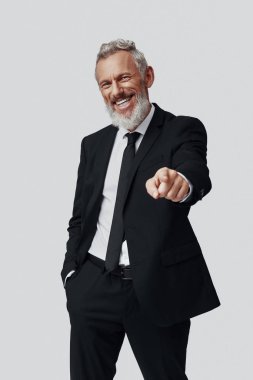 Kendine güvenen, takım elbiseli olgun bir adam gri arka planda dikilirken kameraya bakıyor.