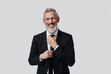 Takım elbiseli, şık, olgun bir adam. Kravatı ayarlıyor ve gri arka planda kameraya bakıyor.