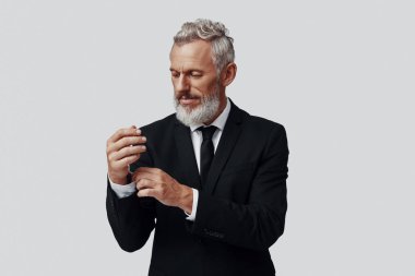 Gri arka planda dikilirken kollarını ayarlayan tam takım elbiseli, zarif, olgun bir adam.