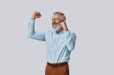 Neşeli olgun bir adam gri arka planda dururken gülümsüyor ve el kol sallıyor.