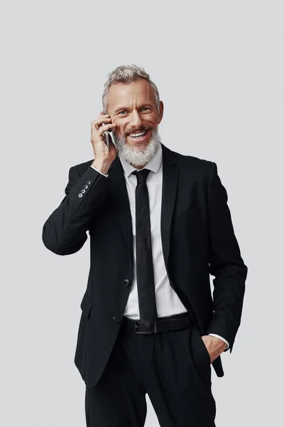 Homem maduro feliz em terno completo falando no telefone e sorrindo enquanto está de pé contra o fundo cinza — Fotografia de Stock