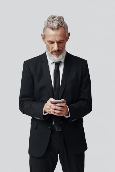 Pensativo homem maduro em terno completo usando telefone inteligente enquanto em pé contra fundo cinza — Fotografia de Stock
