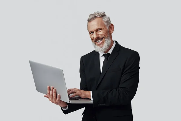 Fiducioso uomo maturo in completo lavoro con computer portatile e sorridente mentre in piedi su sfondo grigio — Foto Stock