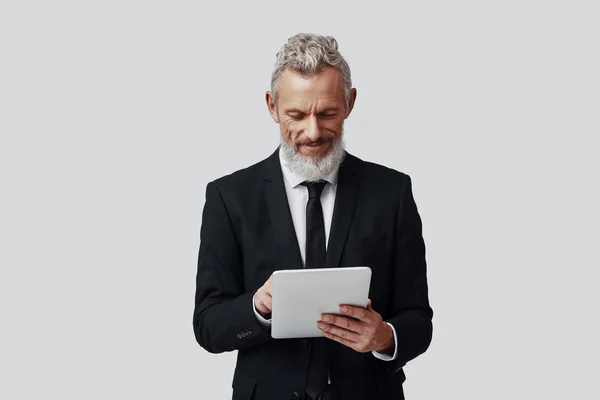 Homem maduro confiante em terno completo trabalhando usando tablet digital e sorrindo enquanto está de pé contra fundo cinza — Fotografia de Stock