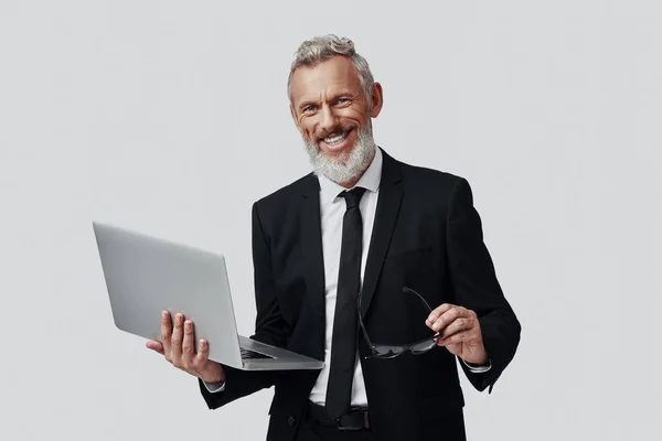 Homem maduro confiante em terno completo trabalhando usando laptop e sorrindo enquanto está de pé contra fundo cinza — Fotografia de Stock