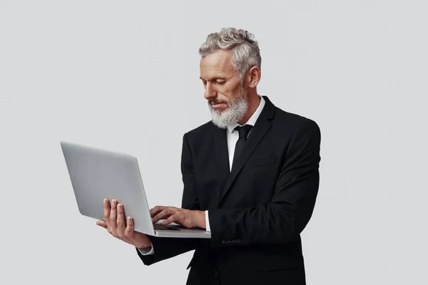 Fiducioso uomo maturo in completo lavoro utilizzando il computer portatile mentre in piedi su sfondo grigio — Foto Stock