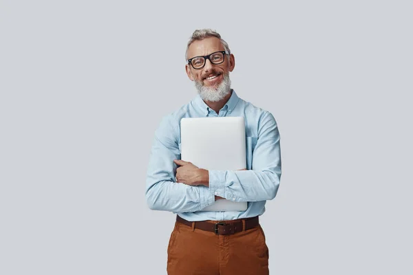 Gelukkig volwassen man glimlachen en dragen laptop terwijl staan tegen grijze achtergrond — Stockfoto