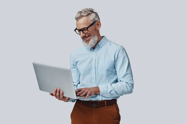 Homem maduro feliz sorrindo e trabalhando usando laptop enquanto está de pé contra fundo cinza — Fotografia de Stock