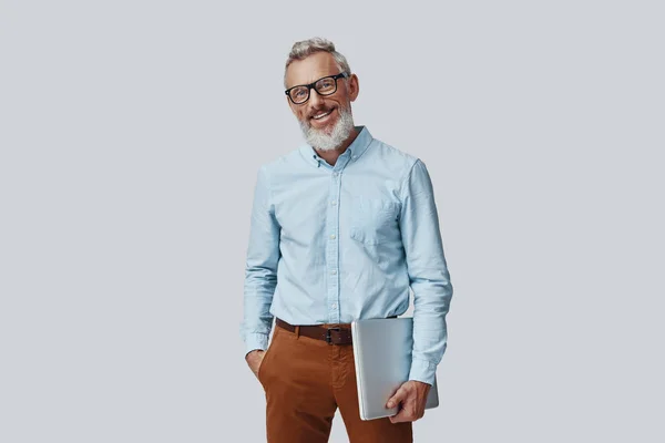 Fiducioso uomo maturo sorridente e portante laptop mentre in piedi su sfondo grigio — Foto Stock