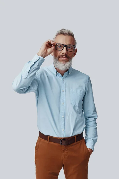 Fiducioso uomo maturo guardando la fotocamera e regolando gli occhiali mentre in piedi sullo sfondo grigio — Foto Stock