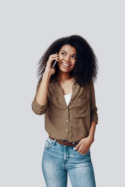 Güzel, genç Afrikalı bir kadın, gri arka planda dururken telefonla konuşuyor ve gülümsüyor. — Stok fotoğraf