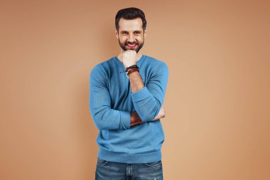Sıradan giyinen yakışıklı genç adam kameraya bakıyor ve kahverengi arka planda dikilirken elini çenesinde tutuyor.
