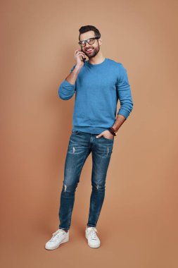 Zeki, rahat giyinen, akıllı telefonla konuşan ve kahverengi arka planda dikilen genç bir adam.