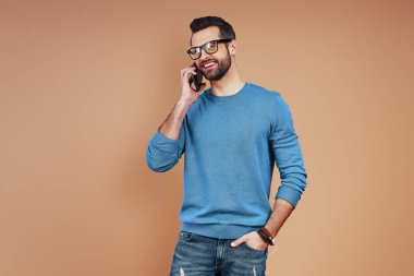 Akıllı gündelik giyinen mutlu genç adam akıllı telefonla konuşuyor ve kahverengi arka planda dururken gülümsüyor.