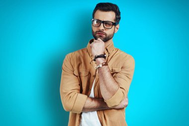 Düşünceli, gündelik giyinmiş genç bir adam mavi arka planda dikilirken başka yere bakıyor ve elini çenesine koyuyor.         