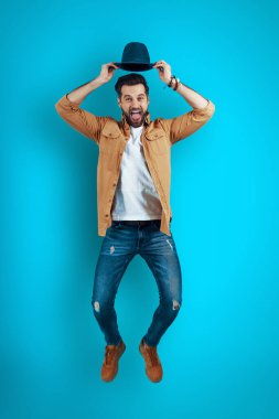 Mavi arka planda süzülürken gülümseyen ve şapka takan yakışıklı bir genç.         