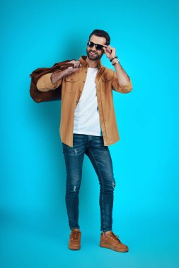 Mavi arka planda dikilirken gülümseyen ve göz giysisini ayarlayan yakışıklı bir genç.