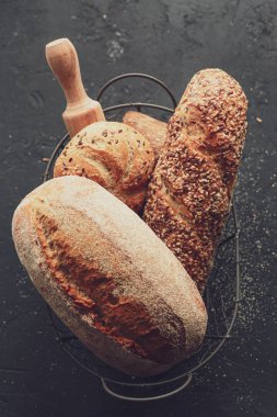 Siyah arka planda sepette ekmek. Metal bir sepetin içinde çeşit çeşit pişirme. Tarif ve metin için yer. Arkaplanda merdane ve un var. Çavdar ekmeği ve ekmekli baget. Karabuğday ekmeği 