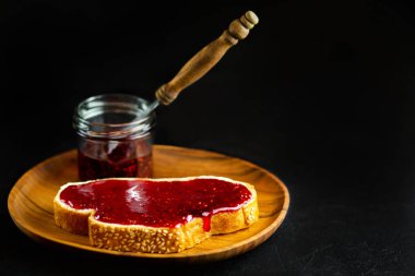 Siyah arka planda reçelli ekmek. Ahşap tabakta beyaz ekmeğe ahududu reçeli. Ahududu reçelli kahvaltı tostu. Vejetaryen kahvaltısı. Vejetaryen yemeği
