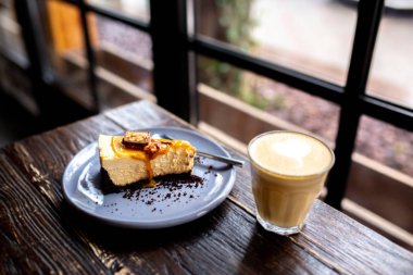 Tahta bir masanın üzerinde bir fincan kahveyle bir dilim cheesecake. Ceviz ve karamel ile cheesecake. Snickers peynirli keki. Karamelli turta. Patlamış mısırlı karamel peynirli kek. Bardakta latte ile tatlı.