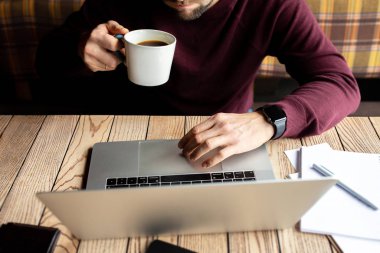 Laptop Bağlantısı Konsepti 'nde çalışan adam. Bu adam çalışıyor. Eller klavyeye yazı yazıyor. İşadamı, elinde bir fincan kahve olan bir kahve dükkanındaki tahta masaya dizilmiş dizüstü bilgisayar, not defteri ve kalem kullanıyor..