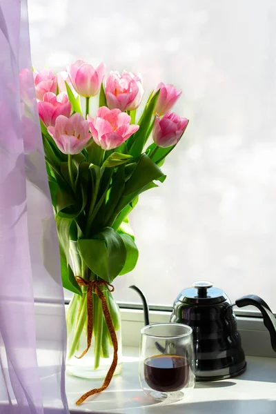 Vase with flowers on the windowsill. A cup of coffee by the window with a bouquet of flowers. Pink tulips in a vase with a teapot. March 8, Women\'s Day. Mother\'s Day. Beautiful romantic morning.