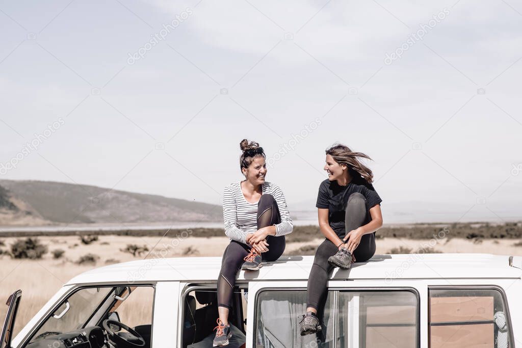 Two happyyoung girls laughing and having fun while sitting on top of the van while traveling. These two girls love adventure, roadtrips and become wild to pursue happiness. Travel Concept. Copyspace