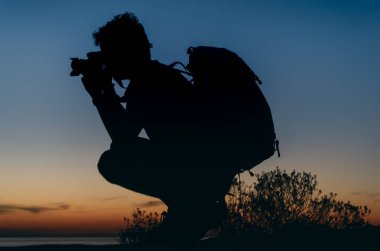 Sırt çantalı genç gezgin gün batımında fotoğraf çekiyor. Renkli gökyüzü, mavi ve turuncu renklerinden dolayı fotoğraf severler için mükemmeldir. Silueti göze çarpıyor. Seyahat konsepti. Kopyalama alanı