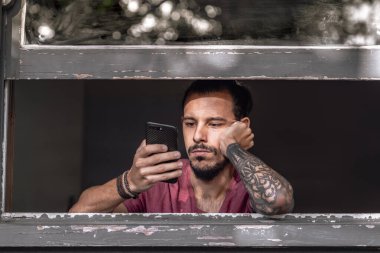 Yüksek dozda akıllı telefondan sıkılan genç bir adam. Koronavirüsün yayılmasını önlemek için güvende olmak için evde kalıyor. Akıllı telefon bağımlılığı var. Konsept: Coronavirus Covid 19