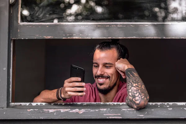 Genç bir adam akıllı telefonunu kullanarak kilitlenmekten zevk alıyor. Koronavirüsün yayılmasını önlemek için güvende olmak için evde kalıyor. O, pencereye yakın oturuyor. Konsept Coronavirus Covid 19. Kopyalama alanı