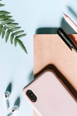 Bir akıllı telefonun, kablosuz kulaklığın, not defterinin ve kalemin dikey düz bir resmi. Pembe çanta mavi arka plan ve yeşil yapraklarla eşleşiyor. Bu modern ve temel çalışma alanı göçebeler ve binyıllar içindir.