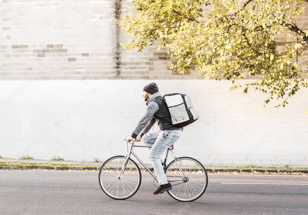Food delivery courier riding a bike on his way to deliver an order. A young and tall male in a silver vintage bike with jeans, beanie and scarf to protect from cold weather. No traffic on the road.