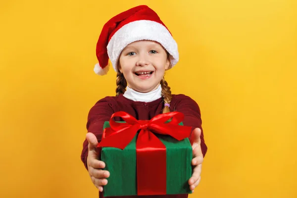 Szczęśliwe dziecko z prezentem świątecznym. Mała dziewczynka w kapeluszu Świętego Mikołaja daje prezent na żółtym tle — Zdjęcie stockowe