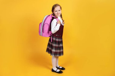 Üniformalı uzun boylu bir kız arkasında sırt çantasıyla sarı bir arka planda duruyor. Sevimli küçük kız gülümsüyor ve kameraya bakıyor.